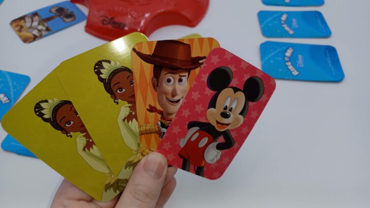 This picture shows a player picking up their first pile in Ring It On!: Disney Edition. They were dealt two yellowish-green Tiana cards, an orange Woody card, and a red Mickey Mouse card.