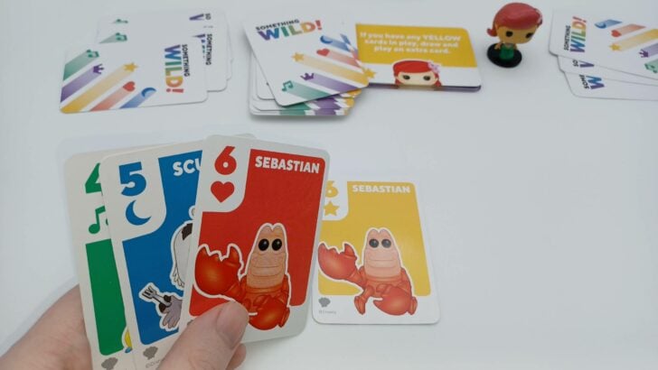 A picture from Something Wild!: The Little Mermaid showing a player's hand with a green four card, blue five, and two Sebastian cards. They have elected to play the yellow Sebastian six card to the table.