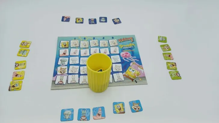 This picture shows how to setup a game of Yahtzee Jr.: SpongeBob SquarePants. The scoreboard is in the middle of the table with the yellow dice cup (and dice inside it) nearby. Each player's tokens are then set up on their side of the play area.